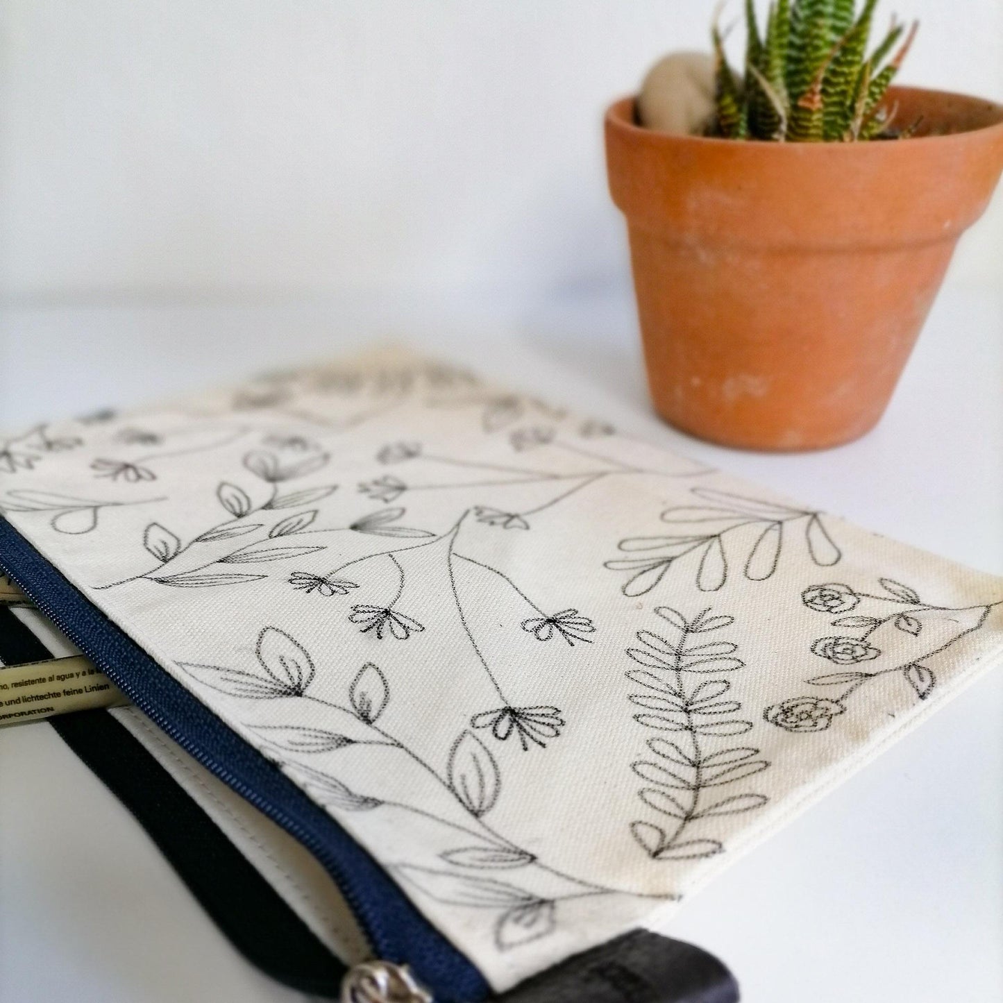 Lined Zipper Cosmetic Bag with hand drawn black veldt flowers. Blue zip. Leather tag. Side view.
