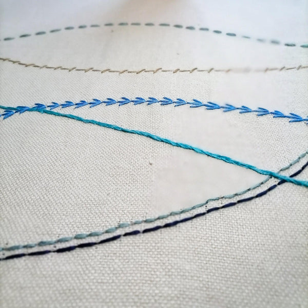 Sand coloured linen table runner with hand embroidered wavy lines in shades of blue. Close up